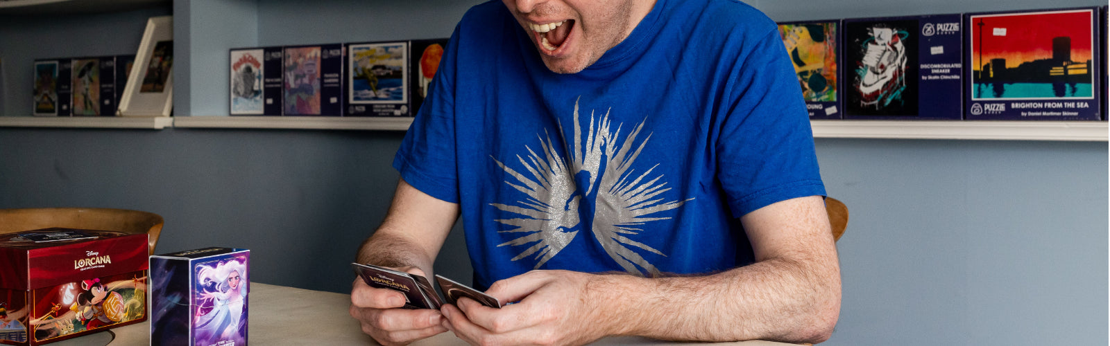 Simon in a blue t-shirt opening Disney Lorcana booster packs and reacting to the cards.